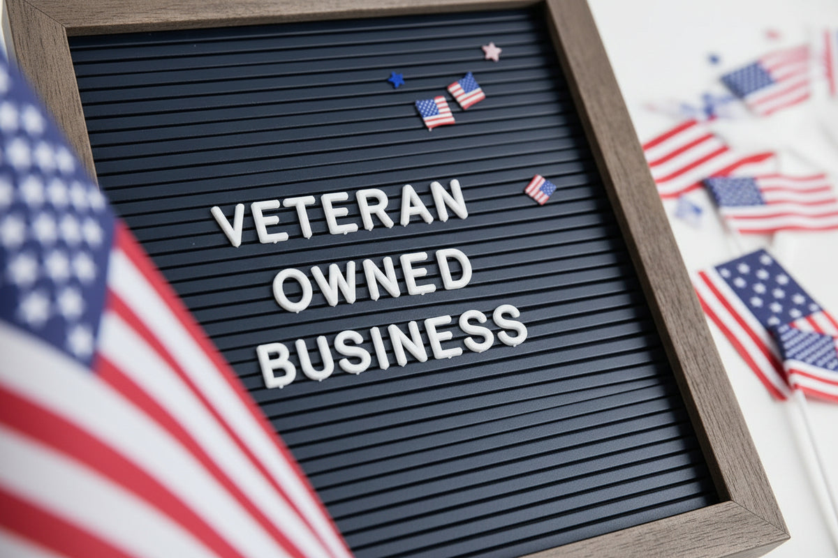 A close-up of a letter board with the words 'VETERAN OWNED BUSINESS' in white letters, decorated with small American flags, with blurred green leaves in the foreground and a patterned surface in the background.